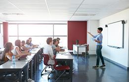 Photo of a teacher and students practising pronunciation in a classroom. E.g.: phonetic chart on the board and teacher pointing at it
