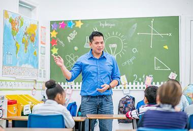 Photo of a teacher and their young students  together in a classroom. Ideally, the teacher  is speaking or writing something on the board.