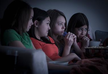 151521 Four female teenage friends watching scary film on sofa with popcorn